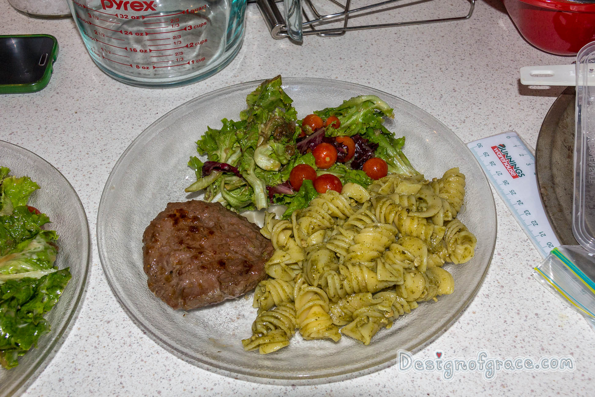 a plate of food with a kangaroo patty on the left which has been nicely browned with pesto pasta on the right and the home grown green and red lettuce and cherry tomatoes on top of the plate