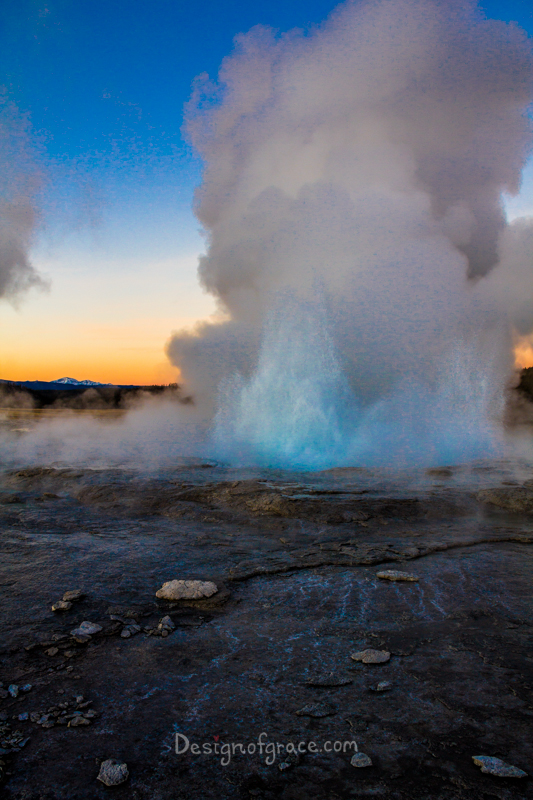 A beauitful orange and blue sunset with a mountain on the left. With the geyser erupting in the front with a blue core. The foreground is dark and rocky