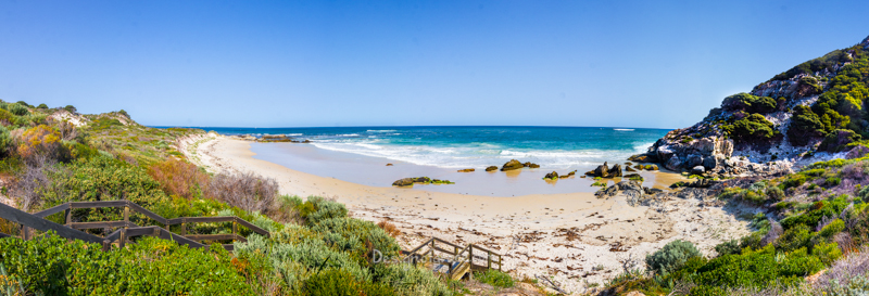 A beautiful panorama of barren's beach. With green bushes on the left with a stairs leading down to the beach. The blue ocean and yellow sand beach in the middle with rocky hills on the right with green bush growing on the hill