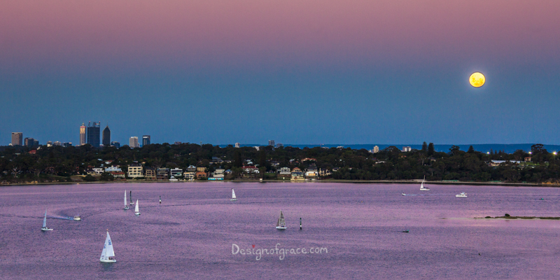 Beautiful purple, pink and blue sunset with the super blue moon on the right above Perth city on the left with yachts sailing in the water in the foreground