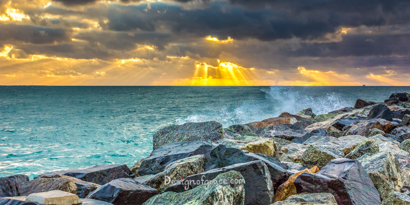 Sunset at Cottesloe over the Indian Ocean with sun rays beaming out of dark and dramatic clouds with colourful rocks on the right
