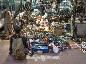 a girl sitting on the bottom left in front of a very full store filled with drills on the floor and chargers on the tables behind the seller