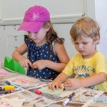 a girl with a pink cap on the left and a boy in a yellow shirt both busy drawing on their bunting